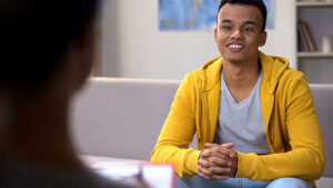a teenager in a yellow hoodie sits on a couch talking to someone about about crown adolescent health and how it can help him